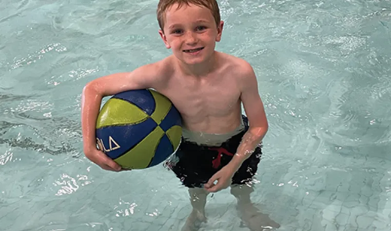 Child in swim trunks standing in a pool holding a ball