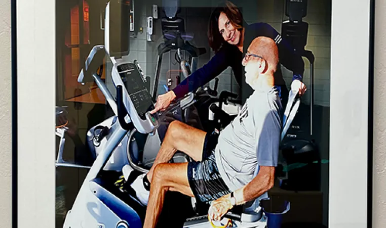 Christine Webb-Miller portrait with her standing next to a man working out on a recumbent bicycle.